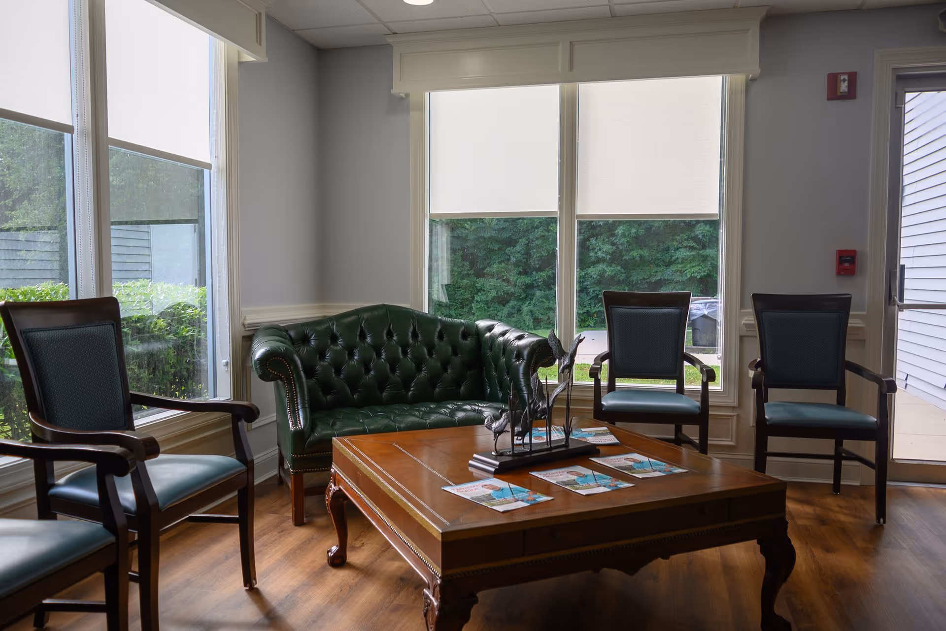 A cozy sitting area with a green leather tufted loveseat, three wooden chairs with blue cushions, and a wooden coffee table with decorative items and brochures on it. Large windows with white roller shades let in natural light and show greenery outside.