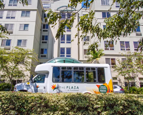 A white shuttle bus parked in front of a multi-story senior living facility building. The bus has the logo and text 'The Plaza at Punchbowl' and 'Senior Living Rental Community' on its side. The building has multiple windows and is surrounded by greenery and trees.