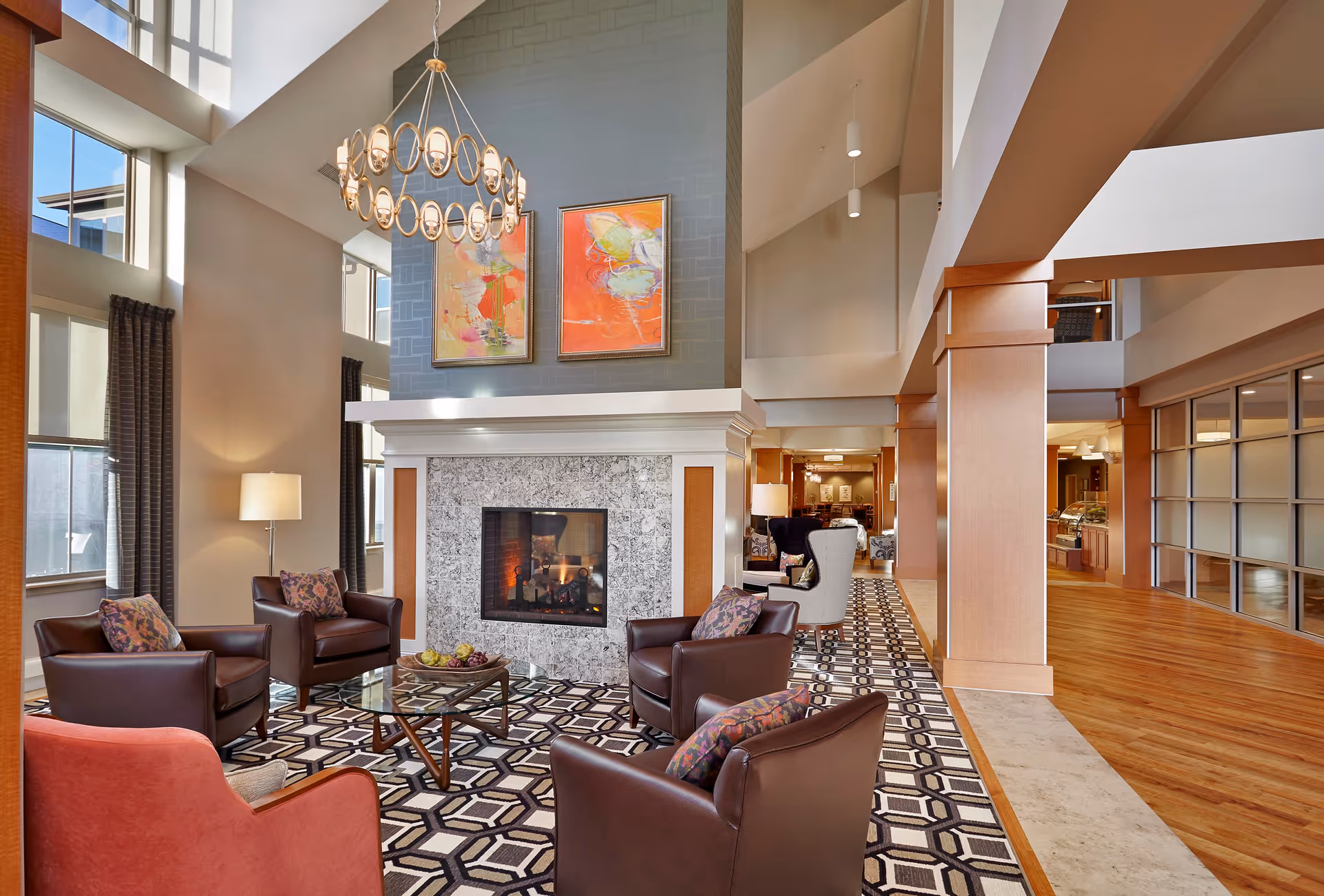 A spacious and well-lit living room area with a modern fireplace surrounded by a marble frame. Four brown leather armchairs and one red armchair are arranged around a glass coffee table on a patterned rug. Two colorful abstract paintings hang above the fireplace. Large windows with dark curtains allow natural light to fill the room. A chandelier with circular lights hangs from the high ceiling. The room extends into a hallway with wooden floors and additional seating areas.