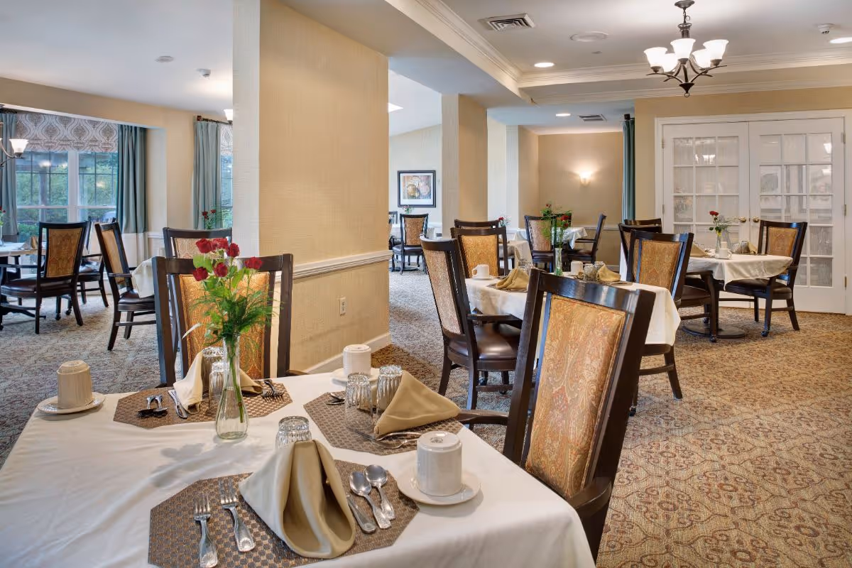 A well-lit dining room in a senior living facility with multiple tables set for meals. Each table has a white tablecloth, beige napkins folded neatly, silverware, cups, and a vase with red flowers. The room features patterned carpet, beige walls, and large windows with curtains allowing natural light to enter. There are wooden chairs with cushioned backs around the tables, and a chandelier hanging from the ceiling.