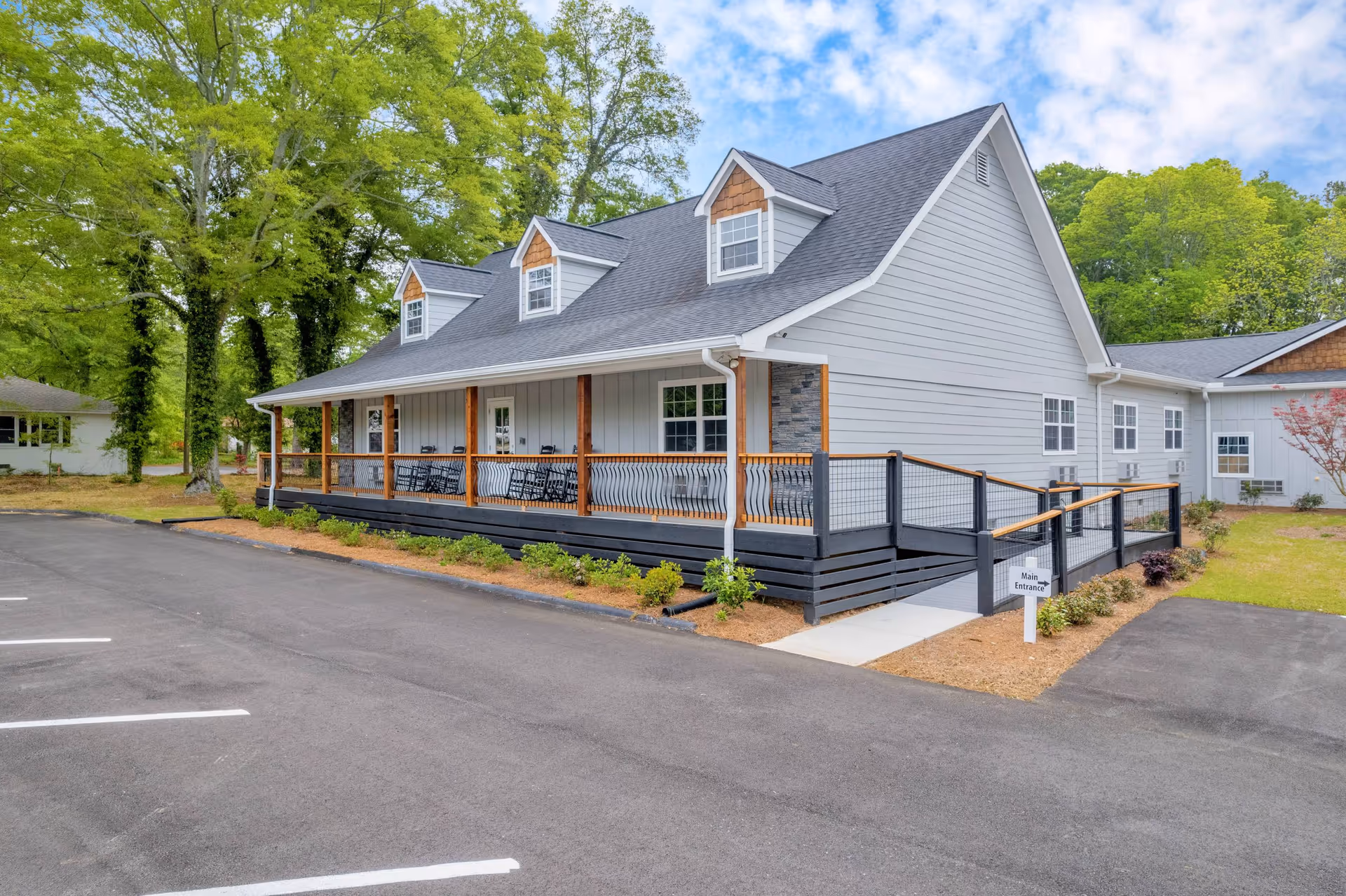 Gray single-story senior living building with a covered front porch, wheelchair ramp, and adjacent parking lot surrounded by trees.