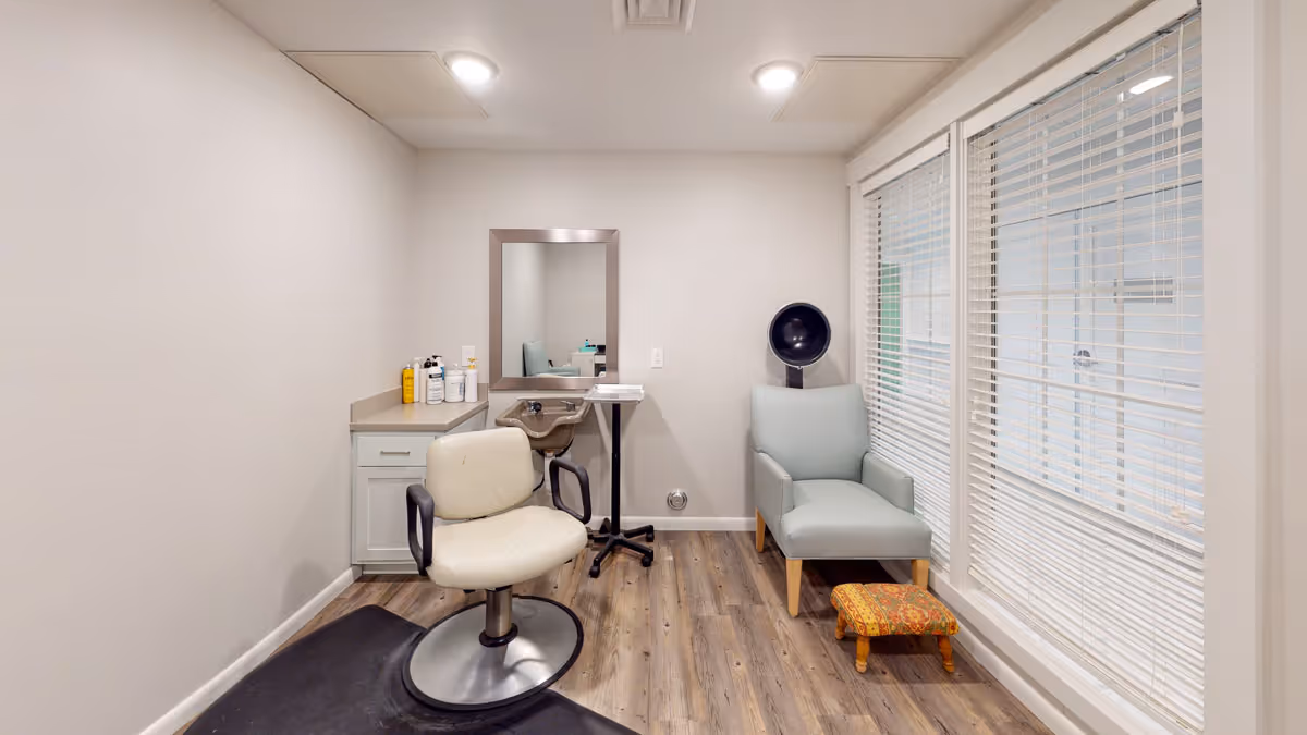 A small hair salon room with a white salon chair on a black mat, a countertop with hair care products, a mirror on the wall, a hair dryer chair, a light blue armchair, a small colorful footstool, and large windows with white blinds.