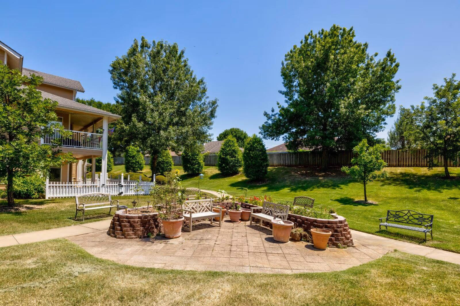 Outdoor patio area with several benches and potted plants arranged on a paved surface, surrounded by green grass, trees, and a two-story building with a balcony in the background under a clear blue sky.