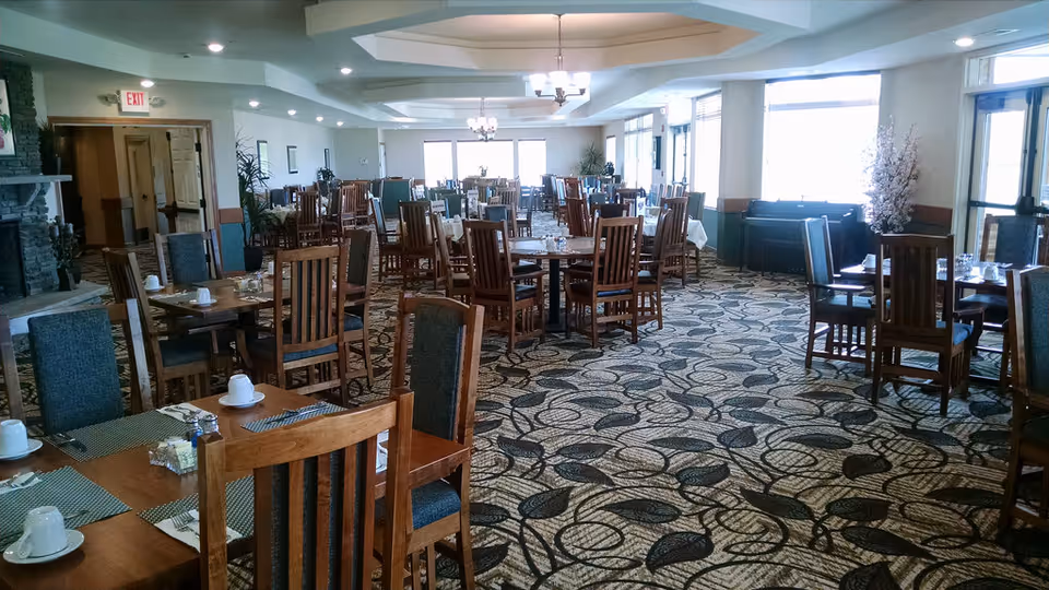Spacious dining room with multiple wooden tables and chairs arranged neatly. Each table is set with placemats, cups, and condiments. Large windows on one side allow natural light to fill the room. The ceiling has recessed lighting and chandeliers, and there is a fireplace on the left side of the room.