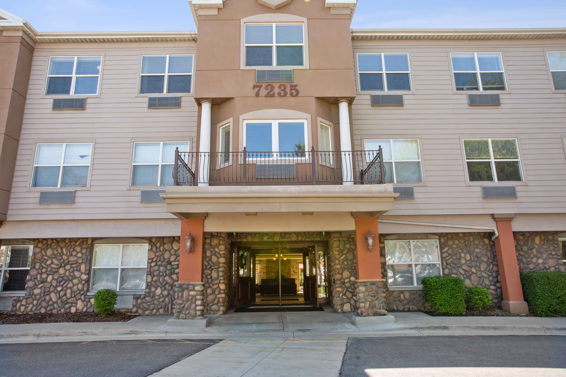 Front exterior view of a multi-story residential building with stone and beige siding, featuring a covered entrance with columns and a balcony above. The building number 7235 is displayed above the entrance.