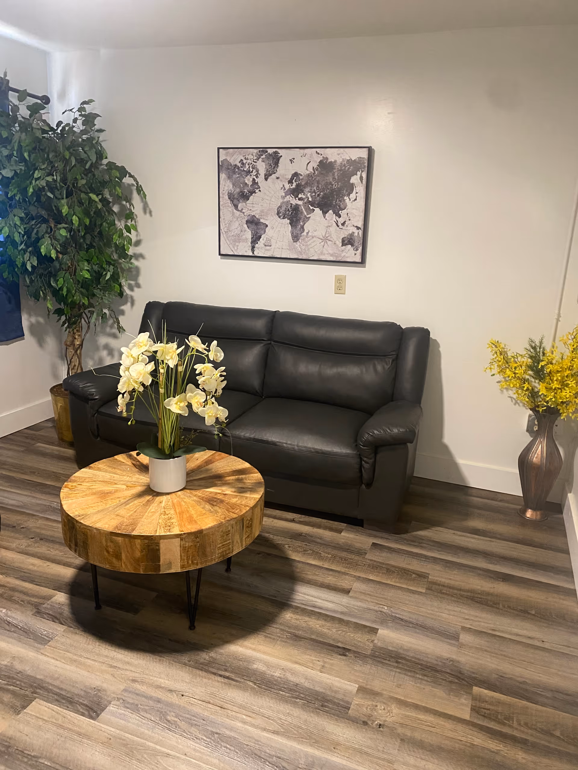 A cozy living room area with a black leather sofa against a white wall. Above the sofa hangs a framed world map. In front of the sofa is a round wooden coffee table with a white vase holding white orchids. To the left of the sofa is a tall green potted plant, and to the right is a tall vase with yellow flowers. The floor is covered with wood-patterned vinyl or laminate flooring.