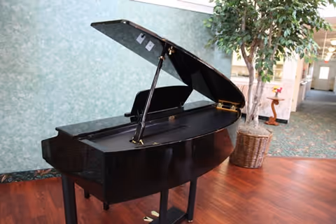 Black grand piano with its lid open in a facility common area beside a potted tree and a hallway.