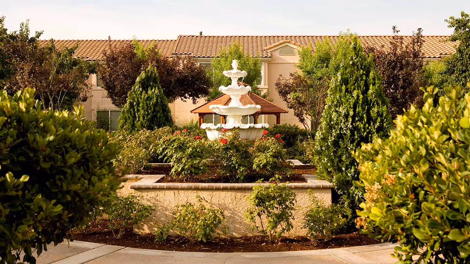 A landscaped courtyard featuring a multi-tiered white fountain surrounded by shrubs and greenery with a building in the background.