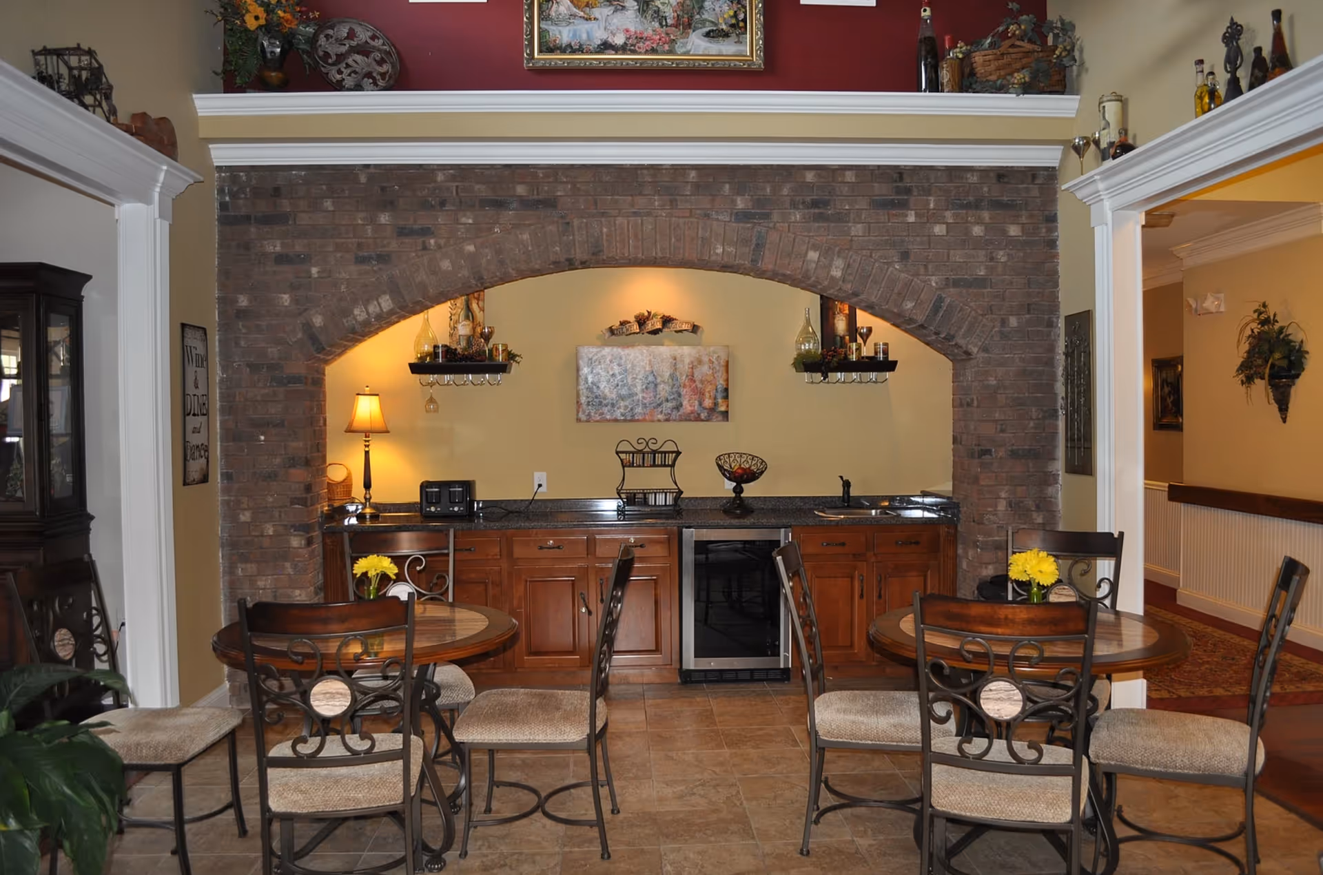 Interior view of a cozy dining area with two round tables, each surrounded by four cushioned chairs with decorative metal backs. The tables have small vases with yellow flowers. Behind the tables is a brick archway framing a kitchenette with wooden cabinets, a small sink, a wine fridge, a toaster, and decorative shelves holding bottles and glasses. The walls are painted beige with a red accent above the arch, and there are various decorative items including a lamp, artwork, and plants.