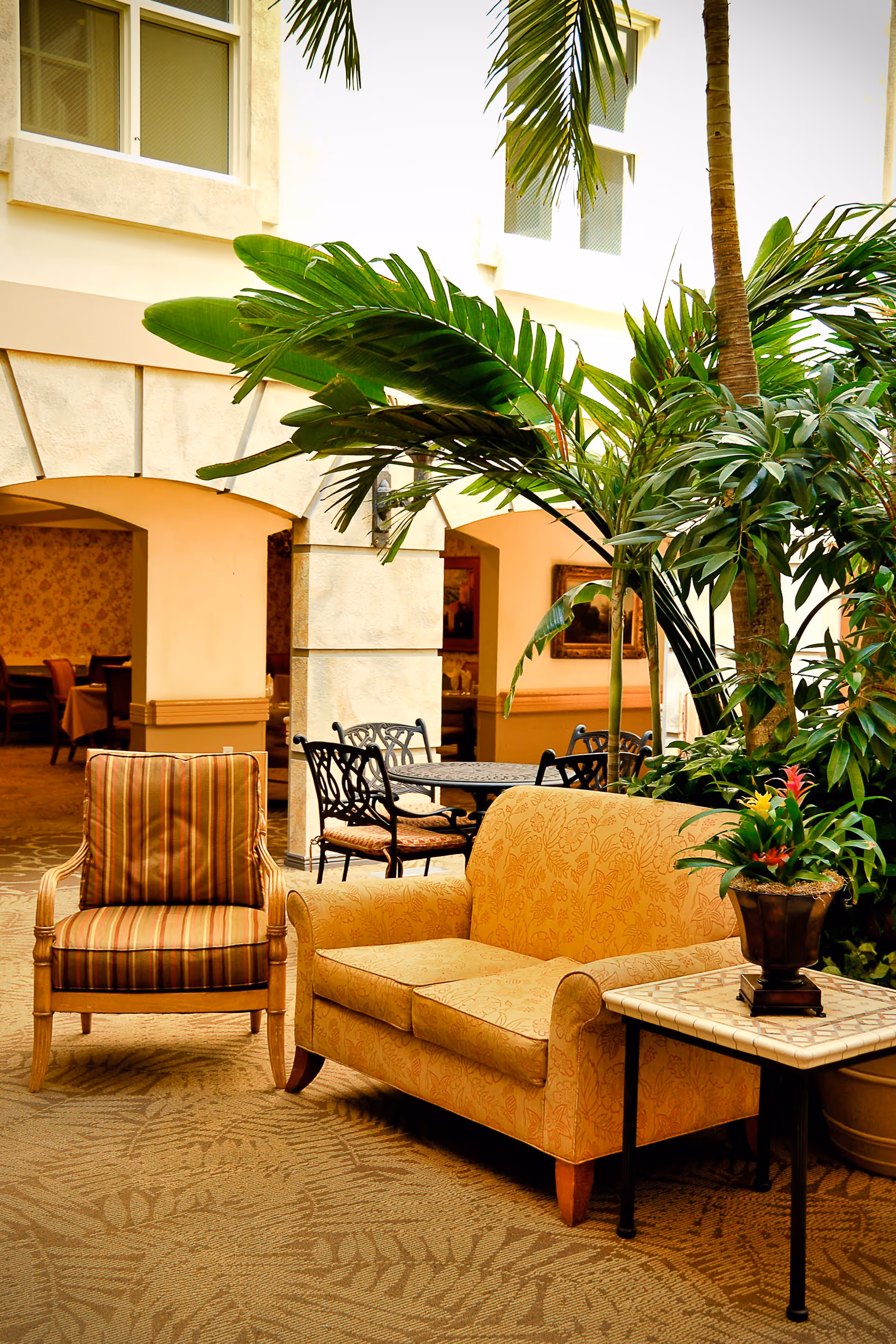Indoor seating area with a patterned yellow loveseat and a striped armchair on a carpeted floor. There is a small table with a potted plant next to the loveseat. Large green tropical plants and palm trees are placed behind the furniture. In the background, there are archways leading to a dining area with tables and chairs.