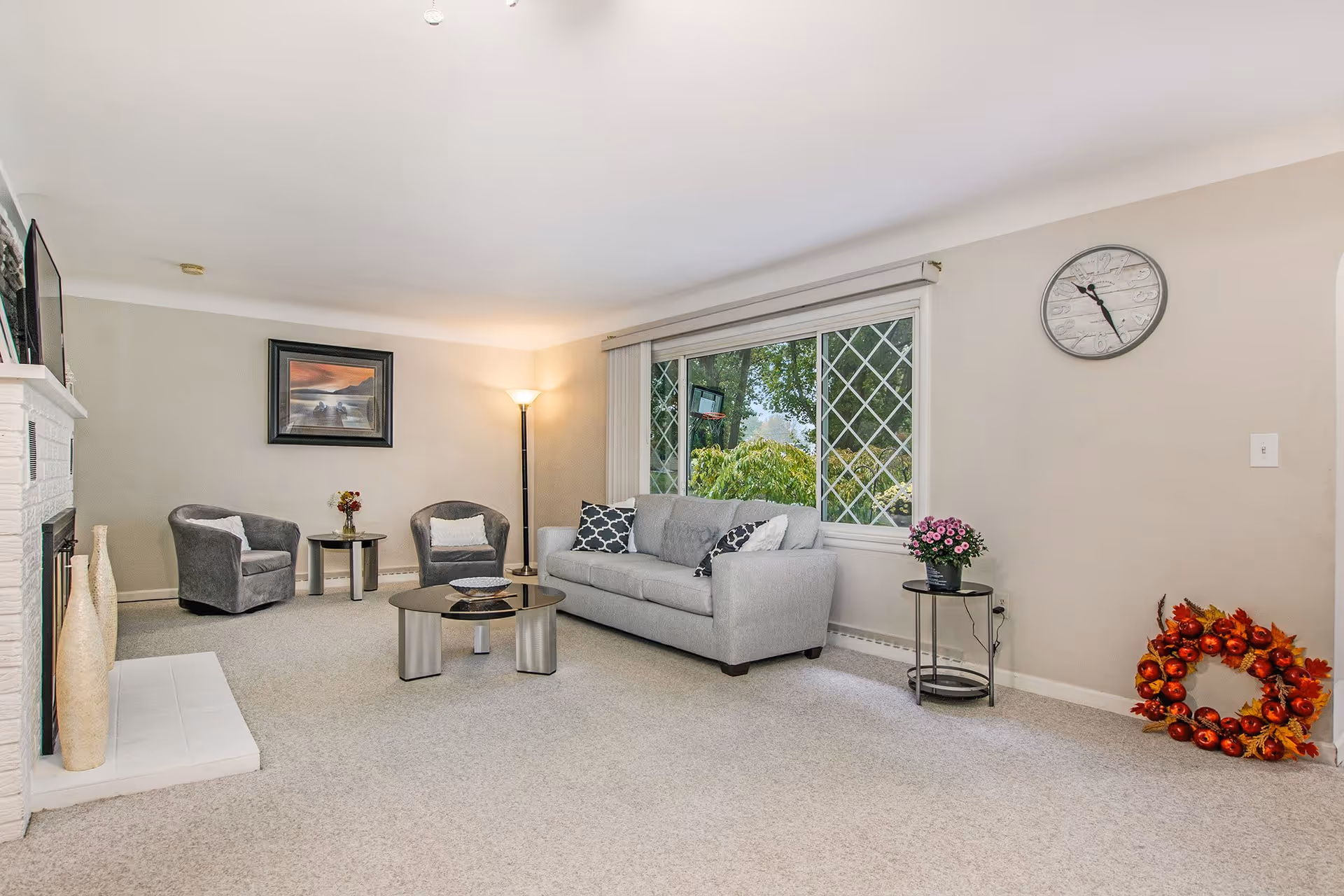 A bright and cozy living room with a light gray sofa adorned with patterned pillows, two gray armchairs, a round glass coffee table, and a side table with a potted plant. There is a large window with diamond-patterned panes letting in natural light, a floor lamp in the corner, a wall clock, a framed picture on the wall, and a decorative wreath on the floor near the wall.
