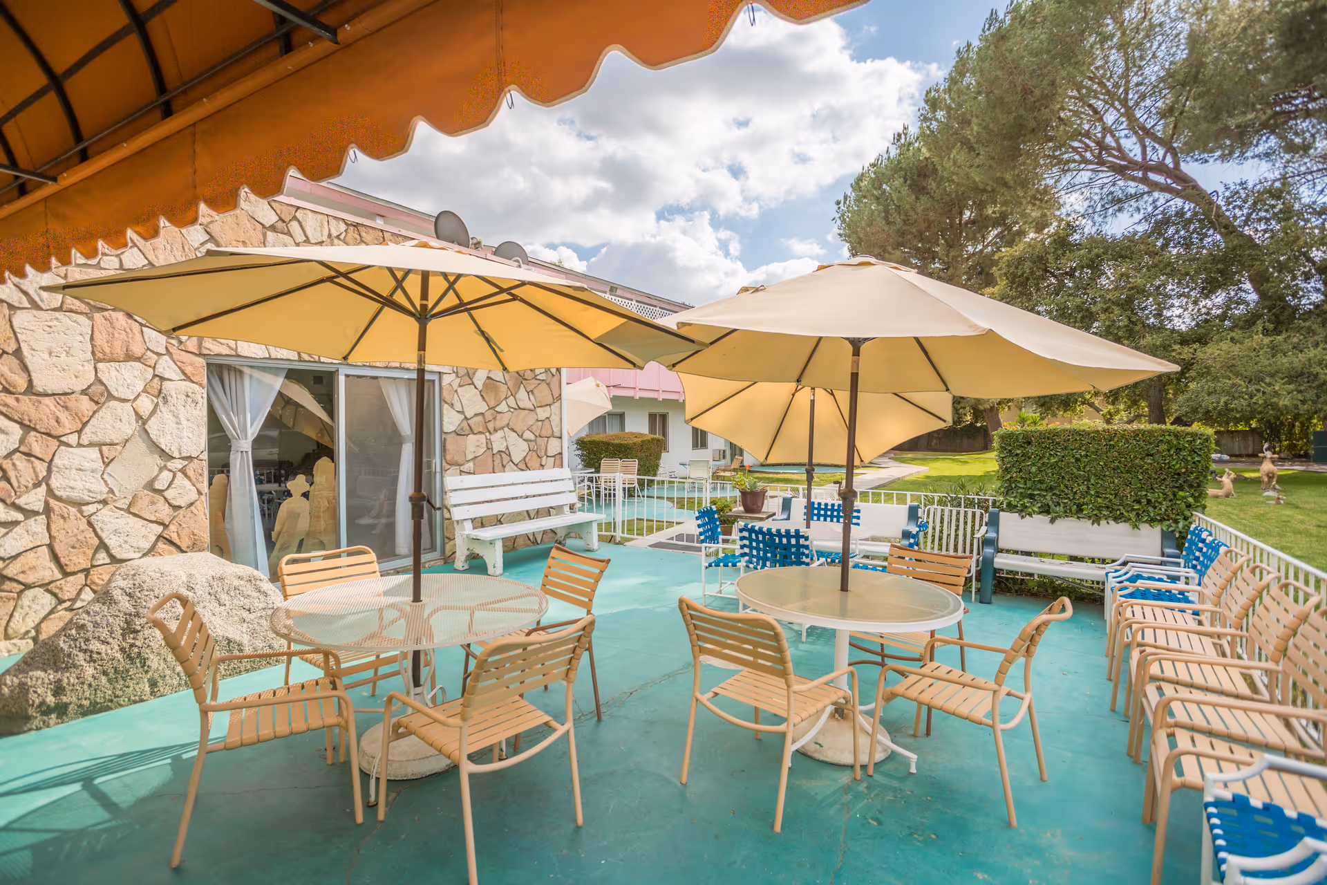 Outdoor patio area with multiple round tables and beige umbrellas providing shade. Each table is surrounded by several beige chairs. The patio floor is painted green, and there are additional benches and chairs along the perimeter. The background shows a stone wall building with large windows and a well-maintained lawn with trees and shrubs under a partly cloudy sky.