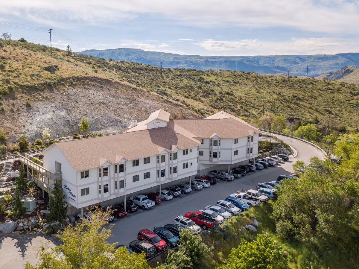 Aerial view of the white multi-story Heritage Heights building with parked cars along the front and hills in the background.