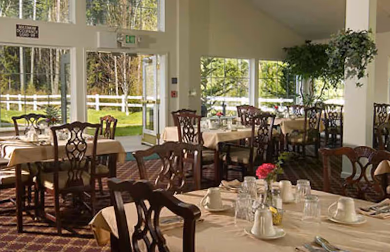 Dining room with multiple tables covered with beige tablecloths, set with cups, glasses, and silverware. Wooden chairs with decorative backs surround the tables. Large windows and glass doors provide a view of a green outdoor area with trees and a white fence. Potted plants hang from the ceiling and add greenery to the room.