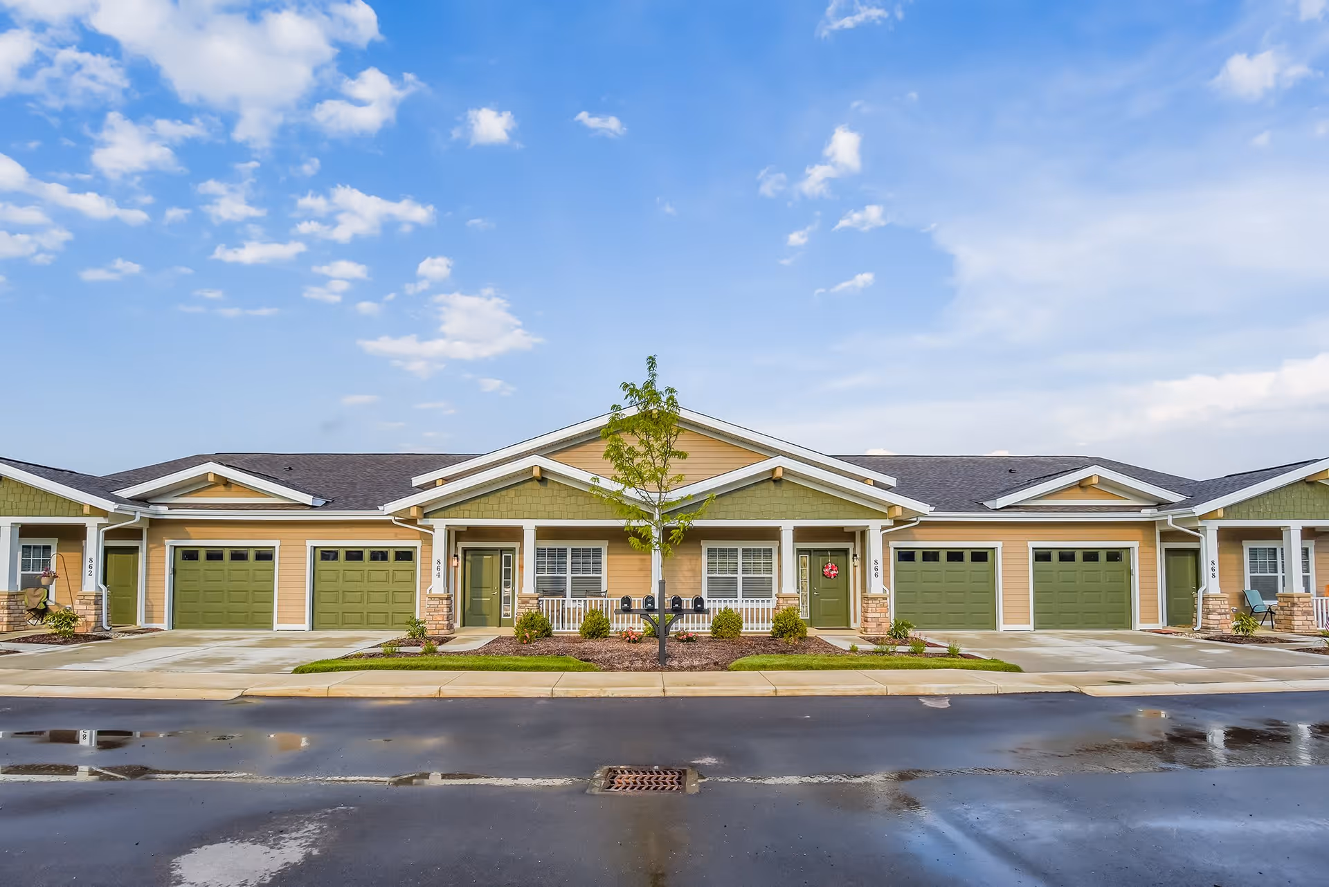 Exterior view of a single-story residential building with multiple attached units, each featuring green garage doors and beige siding. There is a small landscaped area with a young tree and shrubs in front of the building, and a clear blue sky with some clouds above.