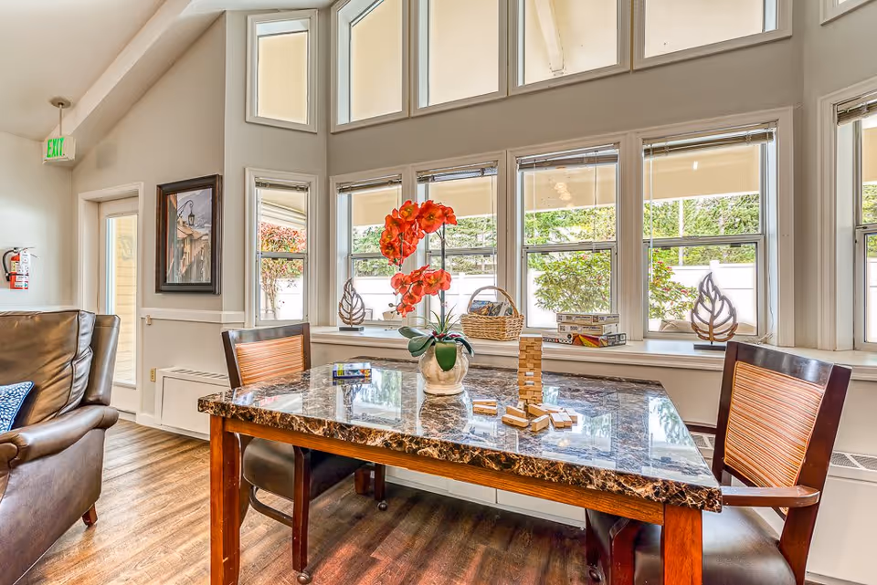 A bright and cozy common area with a marble-top table and two wooden chairs with orange cushions. On the table is a vase with vibrant orange flowers and a partially played Jenga game. Large windows behind the table let in natural light and show greenery outside. To the left, there is a brown leather armchair and a framed painting on the wall near an exit door with an illuminated green exit sign above it.