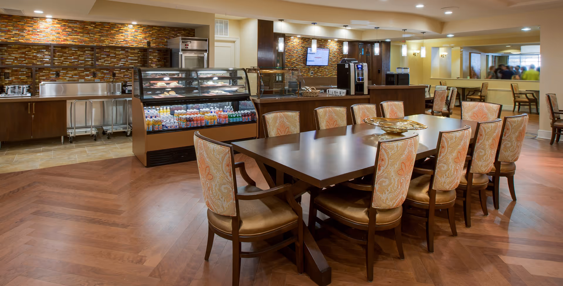 A spacious dining area in a senior living facility featuring a long wooden table surrounded by upholstered chairs with patterned fabric. In the background, there is a food service counter with a glass display case containing beverages and pastries, a coffee machine, and a wall-mounted TV. The room has warm lighting, wood flooring, and a decorative tile backsplash behind the counter.