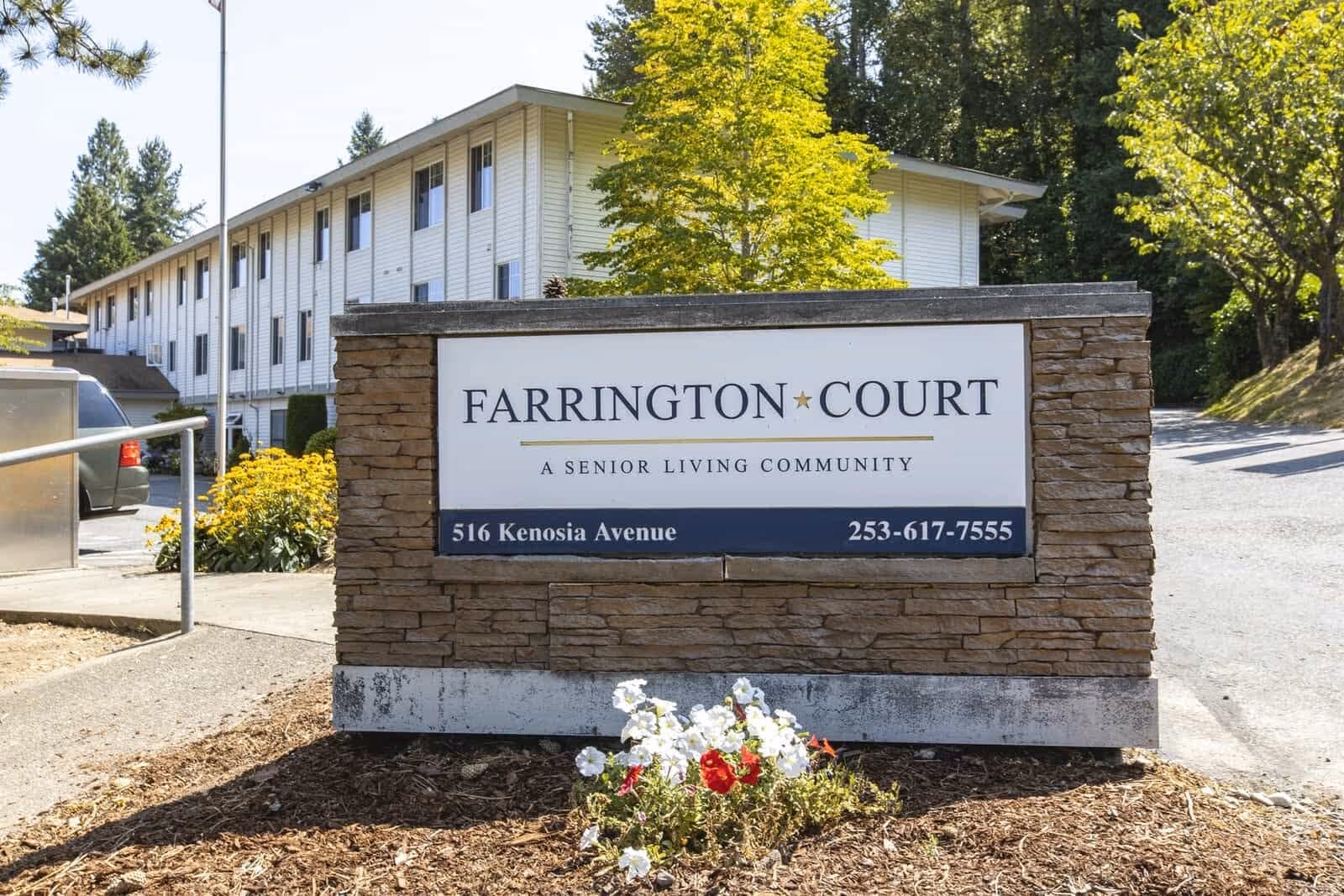 Outdoor view of a senior living community sign that reads 'Farrington Court, A Senior Living Community' with the address 516 Kenosia Avenue and phone number 253-617-7555. Behind the sign is a multi-story building with white siding, surrounded by trees and greenery under a clear sky.