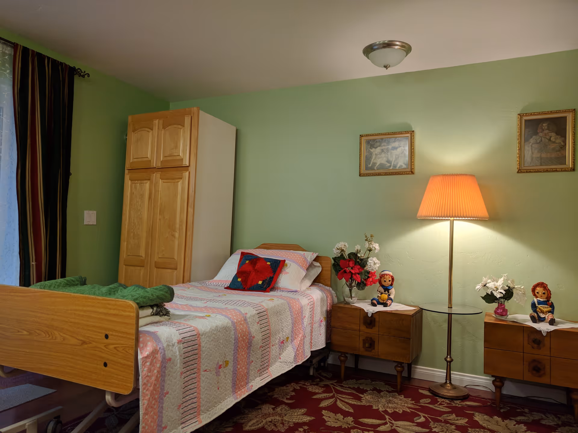 A cozy bedroom with a single wooden bed covered with a pink and white quilt, a green blanket folded at the foot, and a decorative pillow. There are two wooden nightstands on either side of the bed, each with a doll and a vase of flowers. A tall floor lamp with an orange shade is between the nightstands. The walls are painted light green and decorated with two framed pictures. A wooden wardrobe is placed against the wall near the bed, and a window with dark curtains is partially visible on the left.
