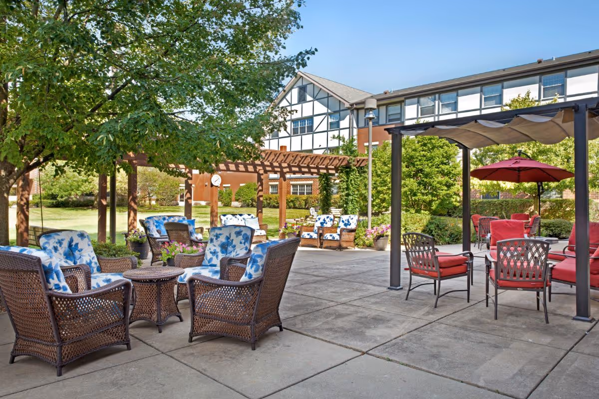 Outdoor patio area at Sunrise of Olympia Fields featuring wicker chairs with blue floral cushions and metal chairs with red cushions under a pergola and umbrella, surrounded by greenery and a building in the background.