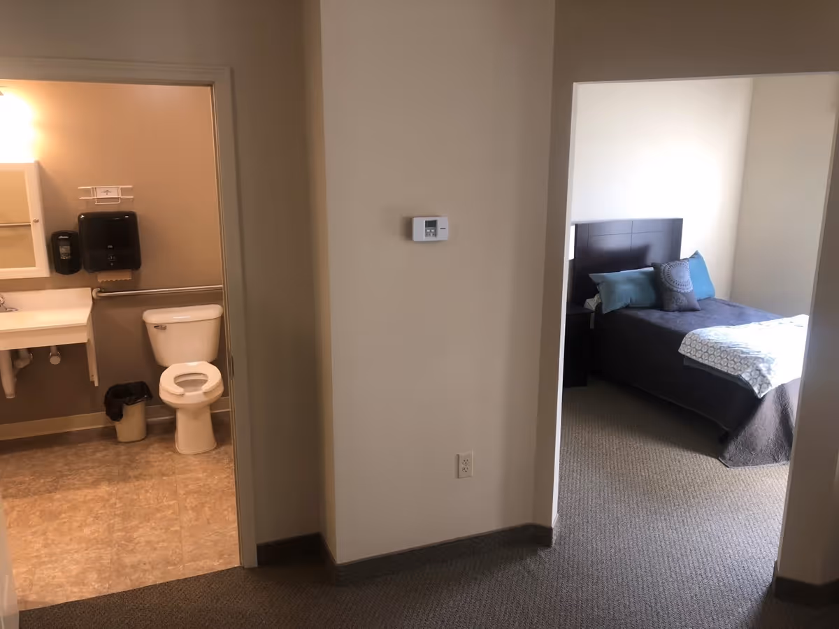 View of a hallway in an assisted living facility showing a bathroom on the left with a toilet, sink, and soap dispensers, and a bedroom on the right with a bed, pillows, and a nightstand.
