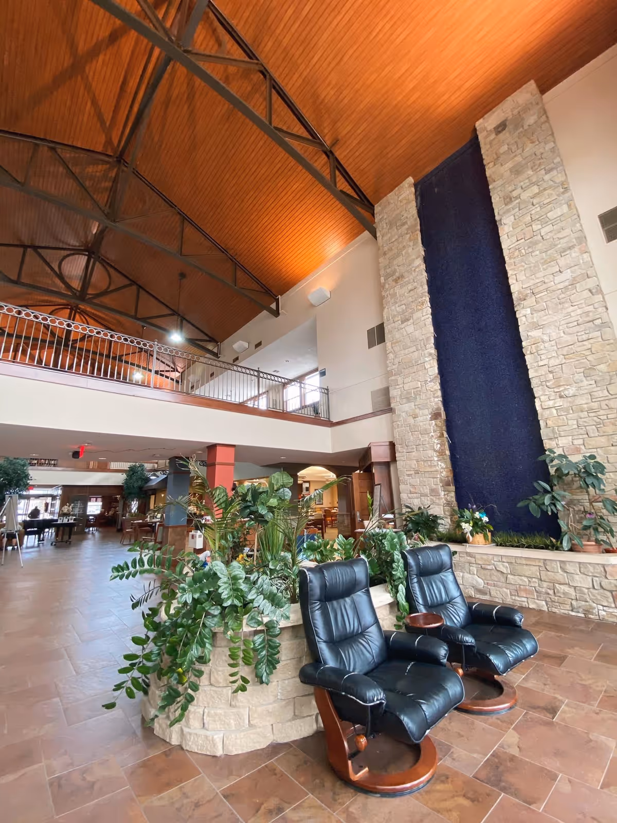 Interior view of a senior living facility with two black leather recliners in the foreground next to a stone planter filled with green plants. The room features a high wooden ceiling with exposed metal beams and a tall stone wall with a vertical blue water feature. In the background, there is a mezzanine level with a railing and various seating and dining areas.