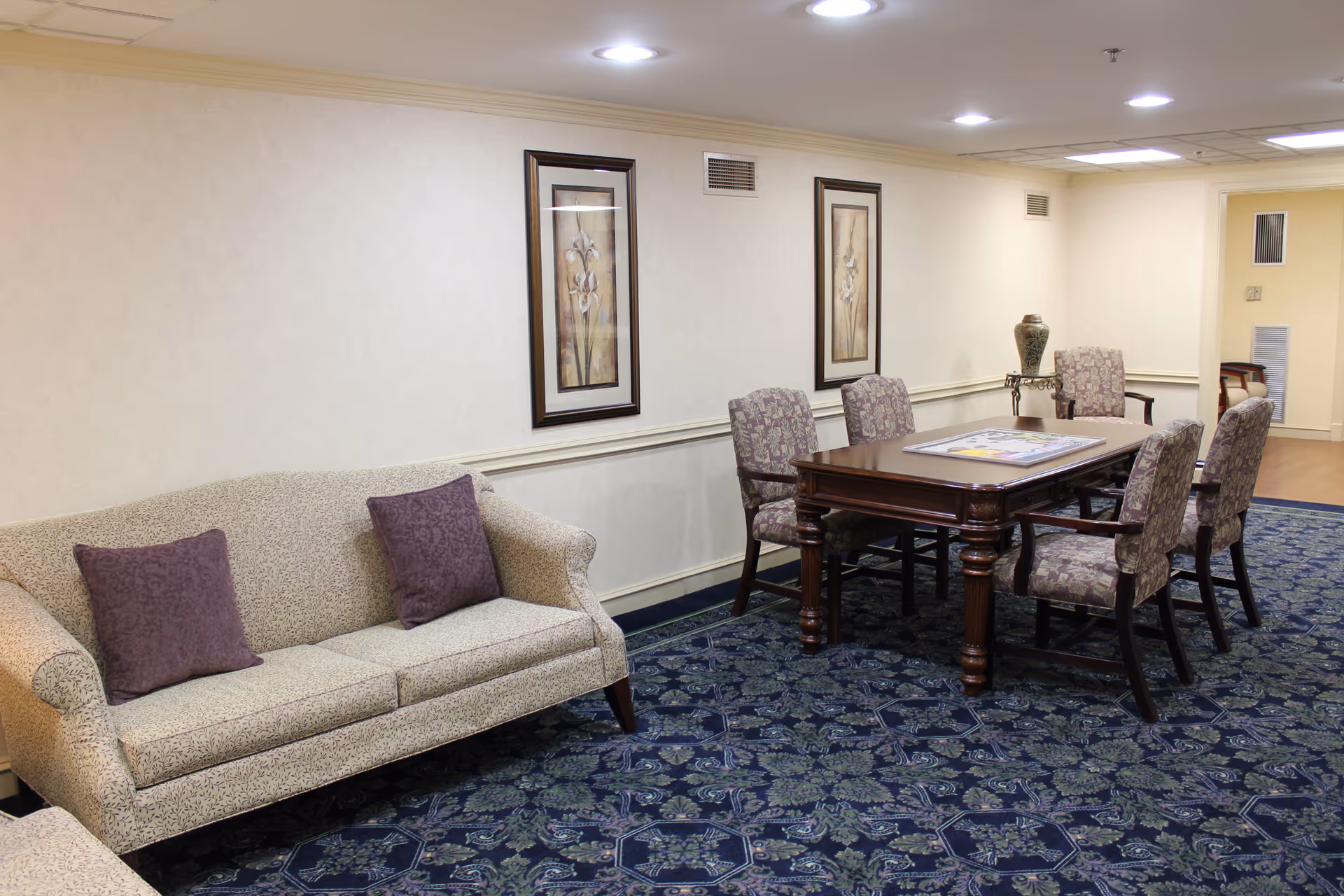 A sitting area in a senior living facility with a beige patterned sofa with two purple cushions, a wooden table surrounded by six upholstered chairs with floral patterns, two framed floral paintings on the wall, and a decorative vase on a small side table. The floor is covered with a blue patterned carpet and the walls are light-colored with white trim.