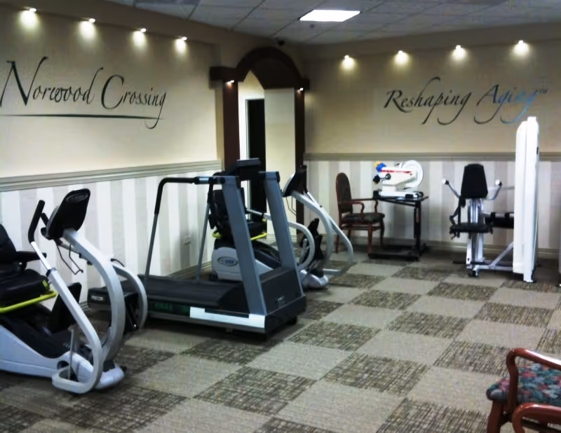 Interior view of a fitness room at Norwood Crossing with exercise equipment including a treadmill, stationary bikes, and strength training machines. The walls have the phrases 'Norwood Crossing' and 'Reshaping Aging' written on them. The room has patterned carpet flooring and recessed ceiling lights.