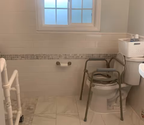 Accessible bathroom with a toilet fitted with a metal commode frame and grab bars, a shower seat, tiled floor, and a frosted window.