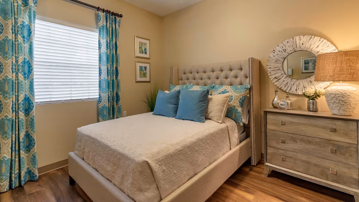 A cozy bedroom with a beige tufted bed frame and a neatly made bed featuring a cream-colored quilt and blue patterned pillows. The room has wooden flooring, a window with blue and beige patterned curtains, two framed pictures on the wall, a wooden dresser with decorative items including a round mirror, a lamp, and a small vase with flowers.