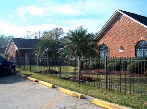 Brick single-story assisted living building with arched windows, a black metal fence, palm trees, and an adjacent parking lot.
