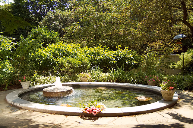 A circular stone fountain with water gently flowing from the center, surrounded by lush green bushes and trees. Several flower pots with colorful flowers are placed around the fountain on a stone-paved area, creating a peaceful garden setting.