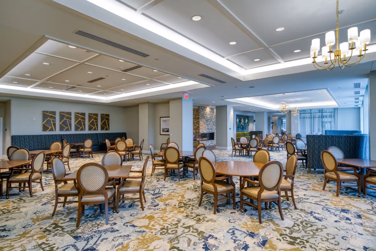 Spacious dining room with multiple round wooden tables and cushioned chairs arranged neatly. The room features a patterned carpet, modern ceiling lights, and decorative wall art. Large windows allow natural light to brighten the space.