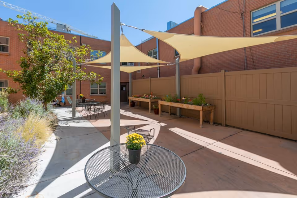 Outdoor courtyard area with metal tables and chairs, some with potted yellow flowers on top. There are shade sails overhead providing partial shade. Raised garden beds with plants are along the fence, and a tree and various plants are visible on the left side. The courtyard is surrounded by brick building walls and a tan privacy fence.