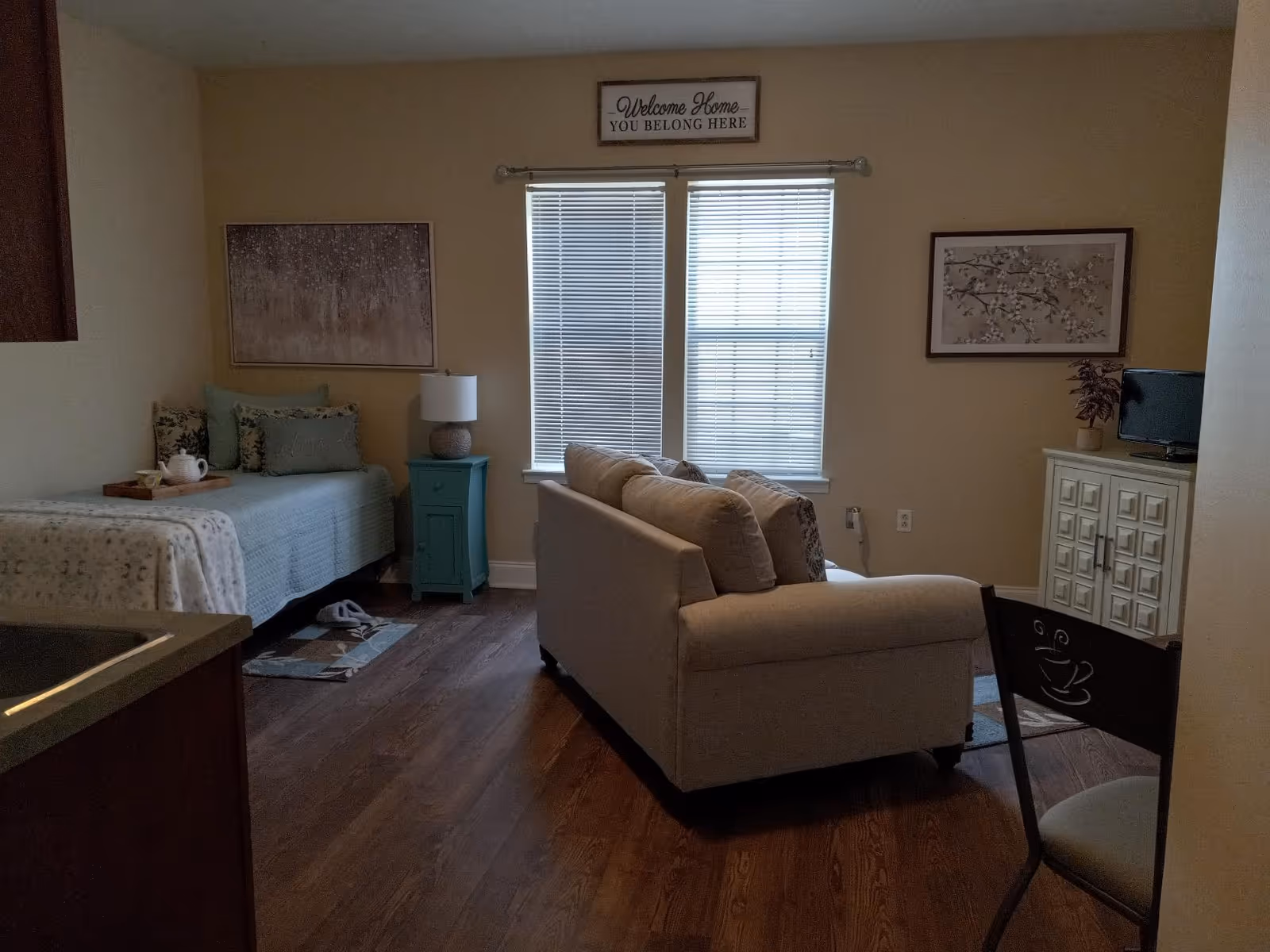 A cozy senior living room with a beige couch facing a small TV on a white cabinet. To the left, there is a single bed with floral pillows and a teal nightstand with a lamp. Two windows with blinds are centered on the back wall, and a sign above them reads 'Welcome Home - You Belong Here'. The room has wooden flooring and warm beige walls.