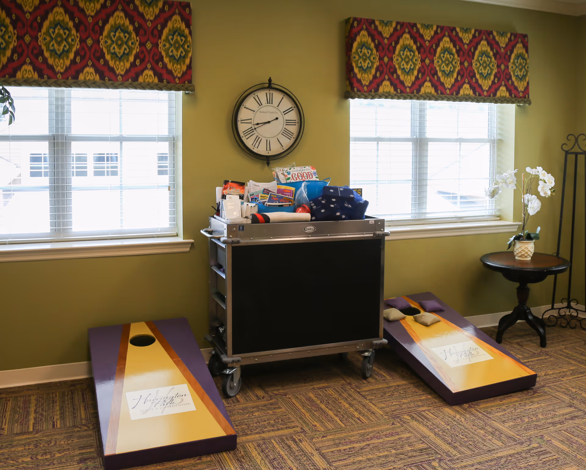 A recreational room with two cornhole boards on a carpeted floor, a cart filled with various games and supplies, a round wooden table with a white orchid plant, two windows with colorful valances, and a wall clock showing the time as 10:10.