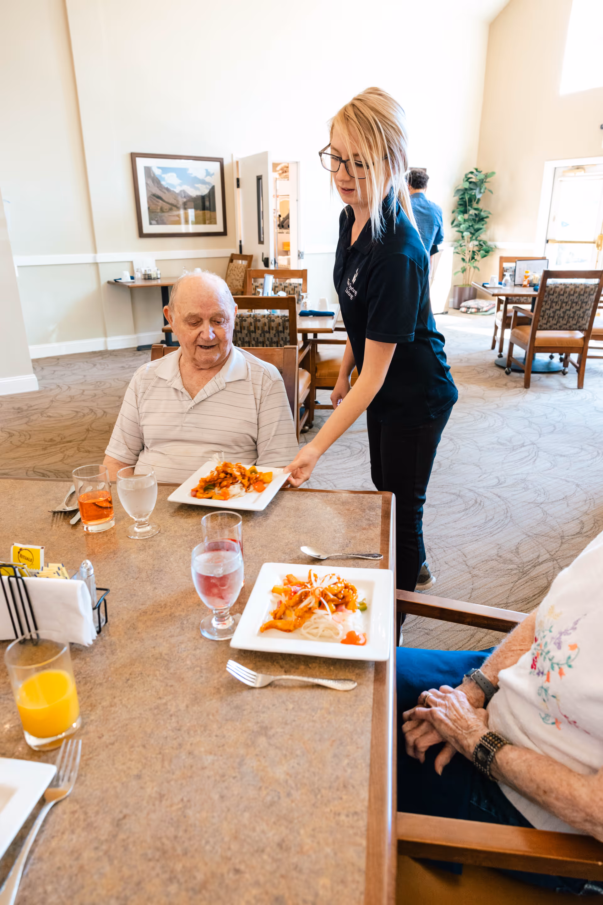 A caregiver serving plated meals to elderly residents seated at a dining table in a bright communal dining room.