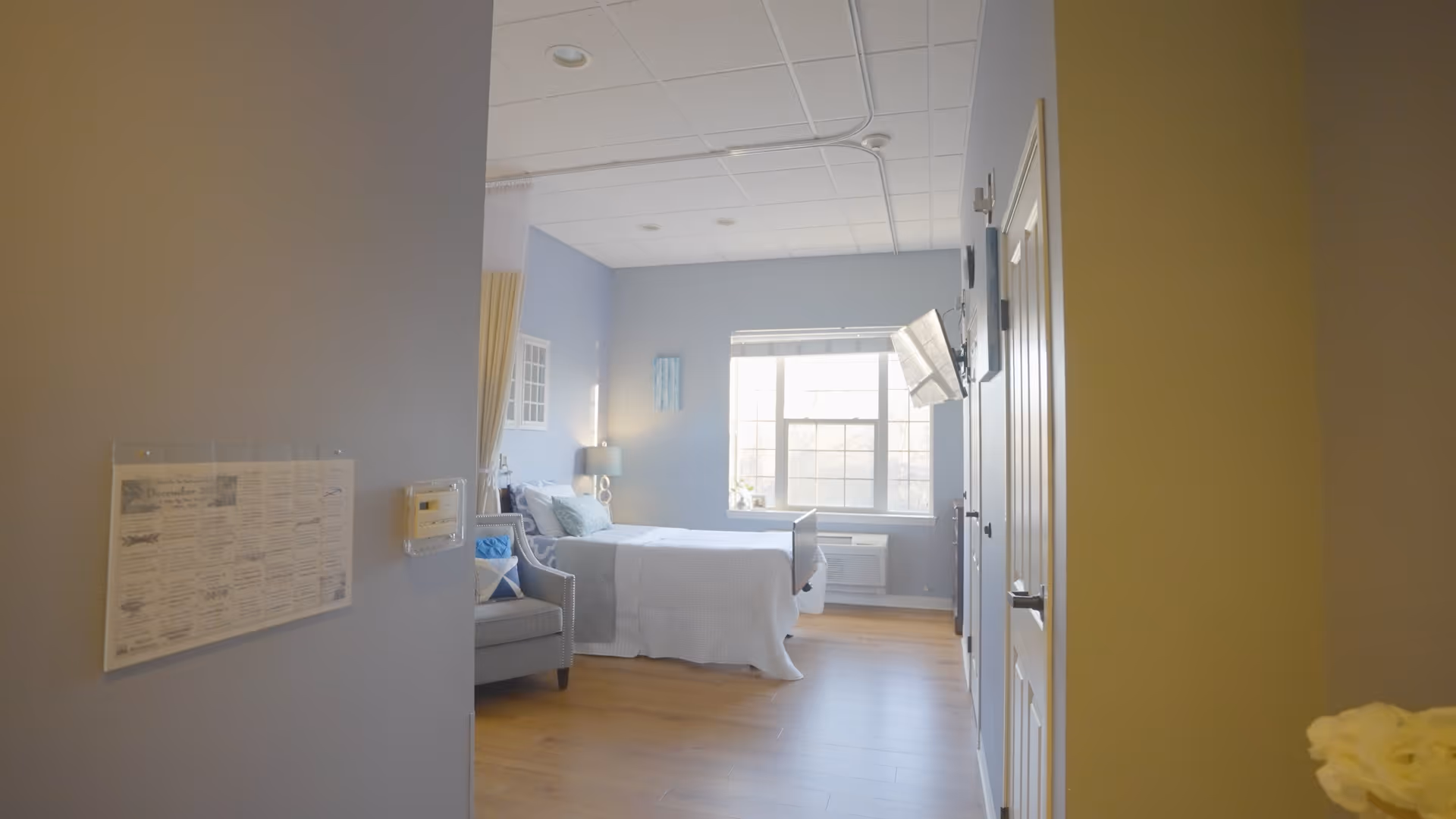 A bright and clean bedroom in a senior living facility with a large window letting in natural light. The room features a neatly made bed with white bedding, a gray armchair with a blue and white pillow, a bedside table with a lamp, and a wall-mounted television. The walls are painted light blue and the floor is wooden.