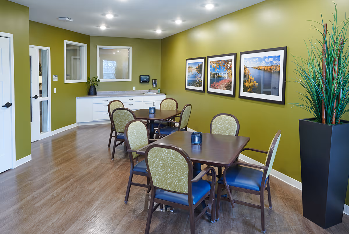 Small dining room with several tables and chairs, green walls, framed landscape photos, and a tall potted plant.