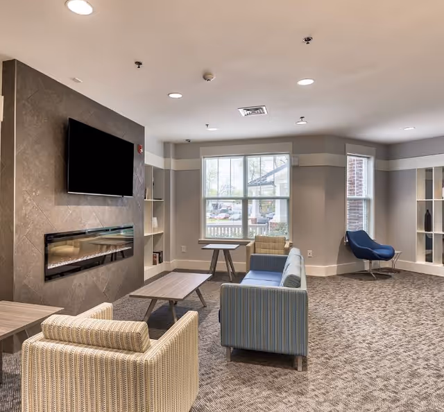 A bright senior living lounge with sofas and armchairs, a wall-mounted TV above a linear fireplace, and large windows.