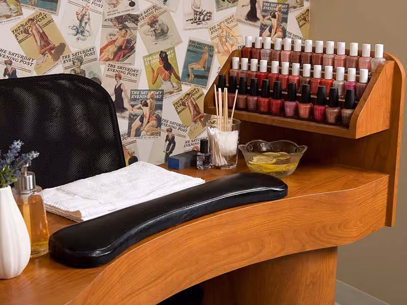 A manicure station with a wooden table and armrest, rows of nail polish bottles, manicure tools and a black mesh chair in front of vintage pin-up wallpaper.