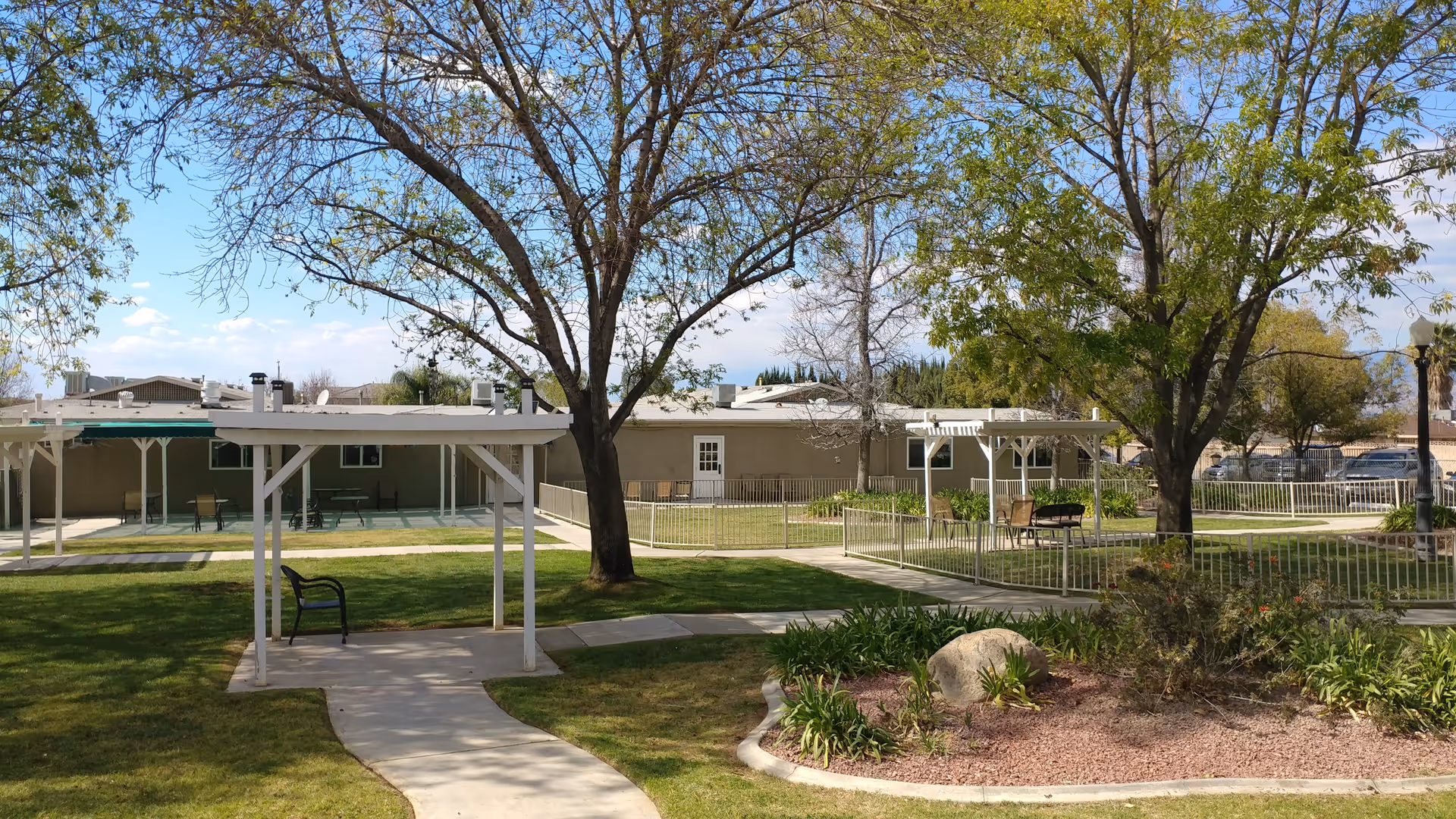 Outdoor garden area at Rose Garden Residential Care featuring green grass, trees with some leaves, paved walkways, white pergolas with benches underneath, and a building in the background under a partly cloudy sky.