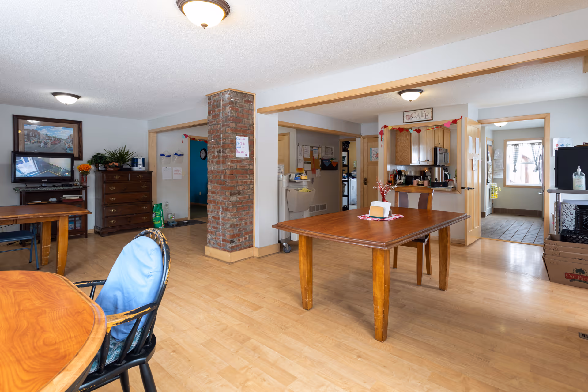 Communal dining/activity room with wooden tables and chairs, a brick column, and a small kitchenette area with a 'CAFÉ' sign.