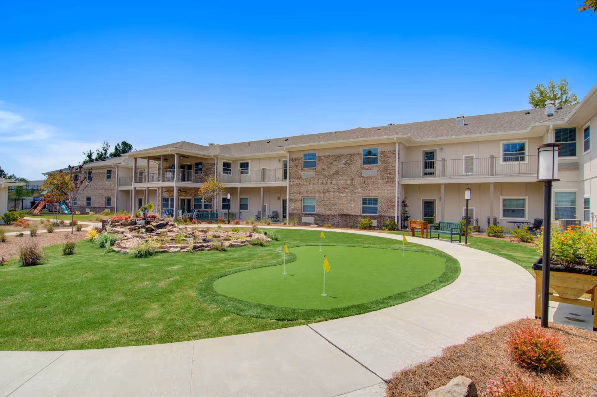 Courtyard of a two-story senior living building with a small putting green, pathways, landscaping and covered balconies.