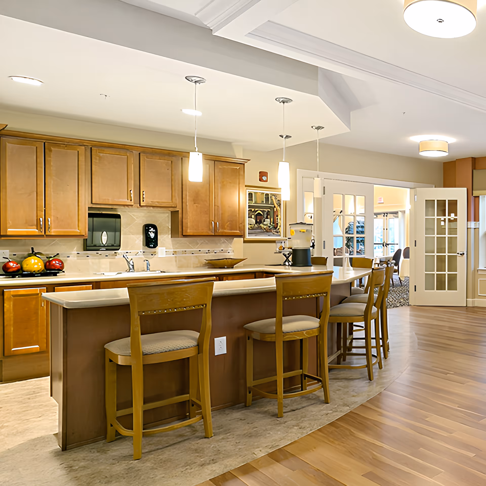 A bright and spacious kitchen area in an assisted living facility featuring a long island with four wooden chairs, wooden cabinets, a sink, and pendant lights hanging from the ceiling. In the background, there are double glass doors leading to another room with seating.
