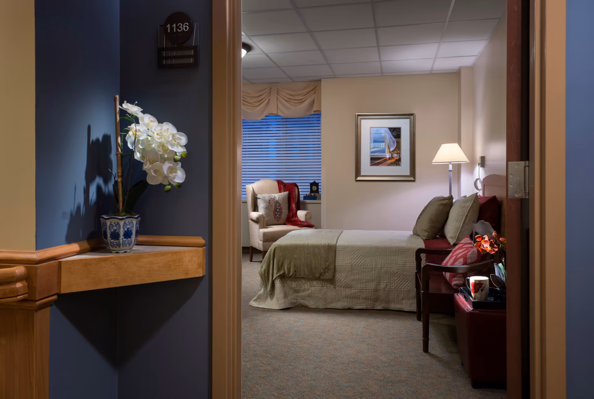 View through a doorway into a furnished senior living bedroom with a bed, armchair, lamp, and decorative flowers on a hallway shelf.