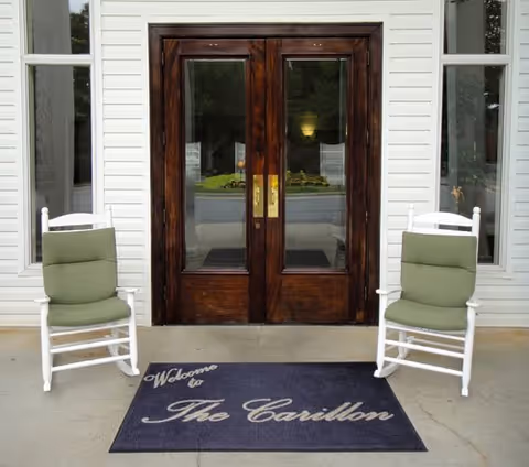 Entrance to a building with double wooden doors featuring glass panels, flanked by two white rocking chairs with green cushions. A dark welcome mat in front of the doors reads 'Welcome to The Carillon'.