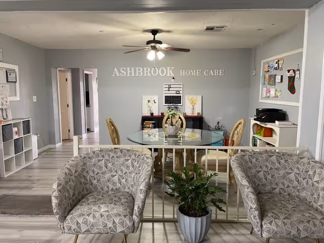 Reception/common area of Ashbrook Home Care with a glass dining table and chairs, two patterned armchairs, a potted plant, and the 'ASHBROOK HOME CARE' sign on the back wall.