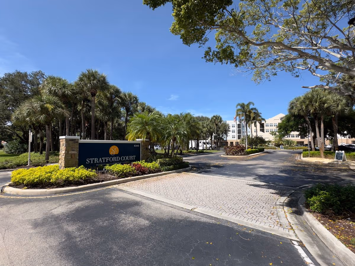 Driveway entrance with a 'Stratford Court' sign and landscaped palms leading to a multi-story residential building.