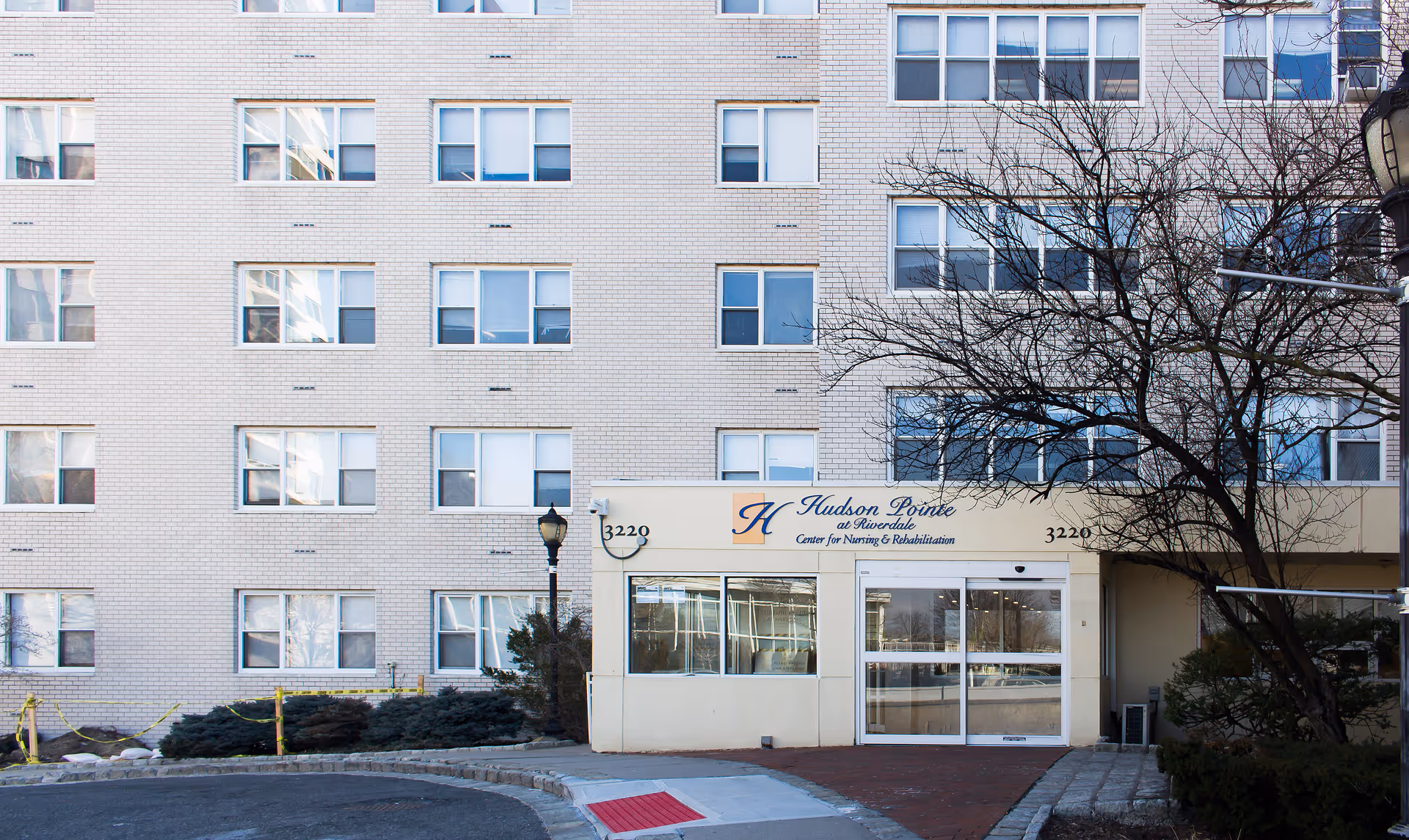 Exterior front view of Hudson Pointe at Riverdale Center for Nursing and Rehabilitation, showing a multi-story brick building with many windows and the main entrance with glass doors and signage above.